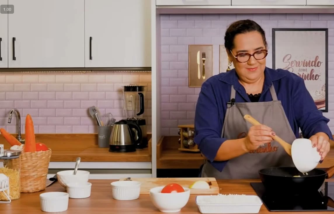 Rosana Silva, de camisa azul e avental, mexendo a panela na cozinha do Servindo com Carinho, com o quadro do programa ao fundo