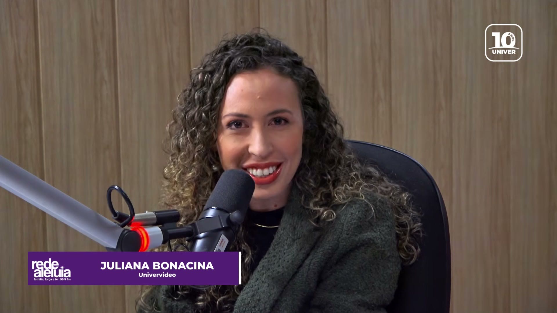 Juliana Bonacina sorrindo ao microfone no estúdio da Rede Aleluia, com o selo dos 10 anos do Univer Vídeo no canto da tela